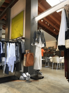 Inside Anastasia Boutique: rails of clothing and a tan leather bag beneath a yellow artwork, with the café counter and white chairs visible in the background under a timber vaulted ceiling
